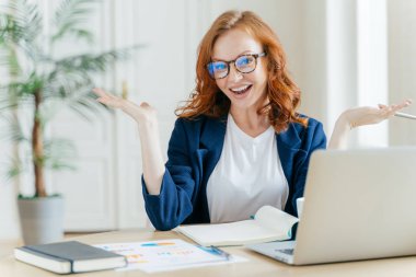 Neşeli Kızıl saçlı işkadını görüntüsünü webinar veya öğretici video izliyor, ücretsiz internet bağlantısı kullanır, avuç içi tereddüt ile yükseltir, masaüstünde kağıtlarla oturur. Küçük işletme sahibi kadın