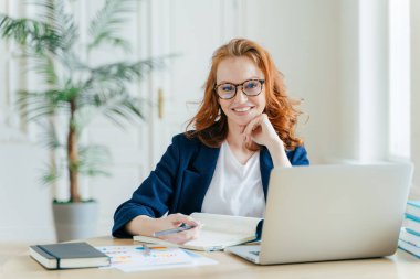 Fotoğraf başarılı zencefil kadın freelancer uzak iş vardır, dizüstü bilgisayar online webinar saatler, yazar bilgileri Not Defteri'nde, mutlulukla gülümsüyor pozlar resmi kıyafetler giymiş işyerinde