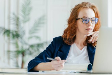 Ciddi deneyimli kadın Koç Kayıt Defteri'nde, dizüstü bilgisayar ekranında odaklı yapar, uzun kızıl saçlı, şeffaf gözlük takar, online webinar izliyor. İş ve iş kavramı