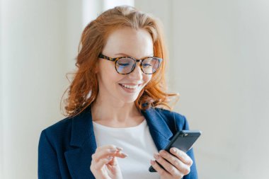Şık giyim, cep telefonu, ileti almak mutlu akıllı pozitif kadın girişimci kar büyütmekten öğrenirse, Kızıl saçlı, gülümseyerek mutlu bir şekilde sahip, modern ofiste kapalı teşkil etmektedir