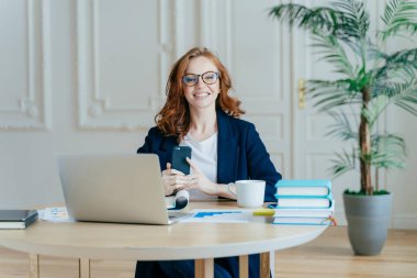 İyimser kadının yatay atış modern cep telefonu uygulama yükler, Kızıl saçlı, resmen giyinmiş bildirim, kağıt belgeleri, dizüstü bilgisayar, eserlerinden ile çevrili olan denetler.