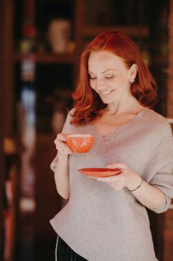 Çekici genç Avrupa Bayan kırmızı kıvırcık saçlı, giymiş gündelik kıyafet, içecek kahve veya çay, boş zaman, gülümsüyor yavaşça, keyfin yerinde olmak, yapmak vardır. Kavram içme insanlar, eğlence,.