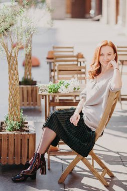 Dikey çekim güzel kırmızı saçlı kadın sandalyede oturur, açık kafe sırada bekler, jumper, etek ve ayakkabı giyer, kameraya memnun ifade ile görünür. İnsanlar, dinlenme ve yaşam tarzı kavramı