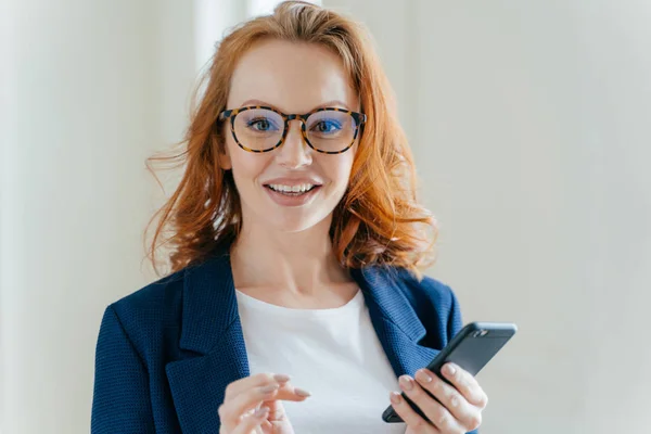 快適な外観と 赤い髪を持つ女性銀行のヘッド ショットの携帯電話オンライン銀行は 金融のウェブサイトに通知をチェック 透明ガラスとエレガントなフォーマルなスーツを身に着けています — ストック写真