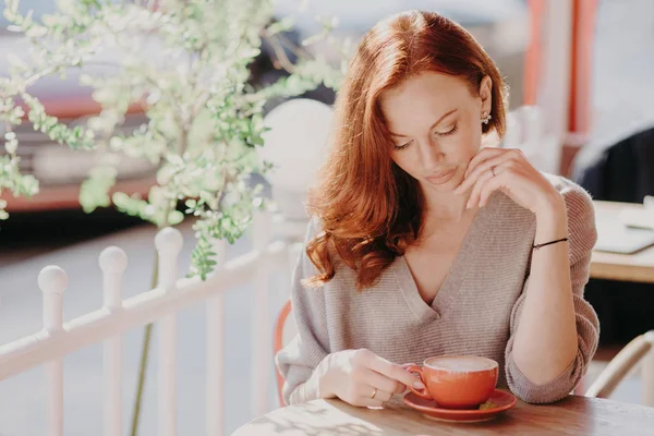 Sevimli kırmızı saçlı Avrupa kadın içecekler cappuccino veya teras cafe kahve, sakin yüz ifadesi olan, kahverengi Kazak giyer, yapmak vardır, iyi içki sahiptir. İnsanlar ve yaşam tarzı kavramı.