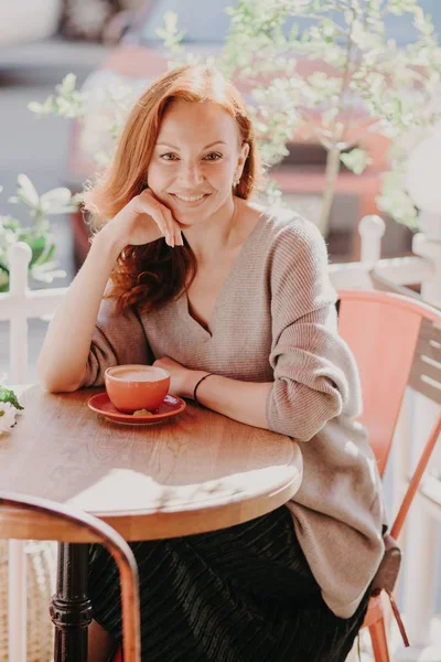 Vertical shot of cheerful foxy female dressed in fashionable clothes, drinks coffee, poses in outdoor cafe, enjoys spare time, has pleasant smile on face, keeps hand under chin. Lifestyle concept