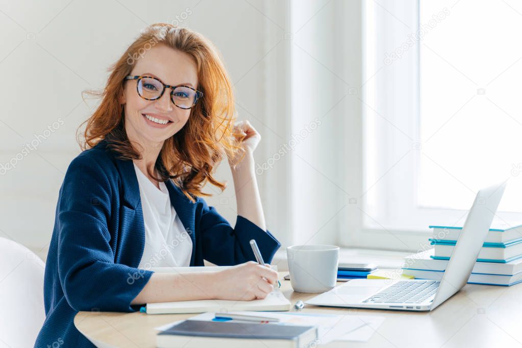 Sideways shot of prosperous businesswoman with foxy hair, smiles positively, records information in notepad, drinks coffee, searches information on laptop computer, develops strartup project