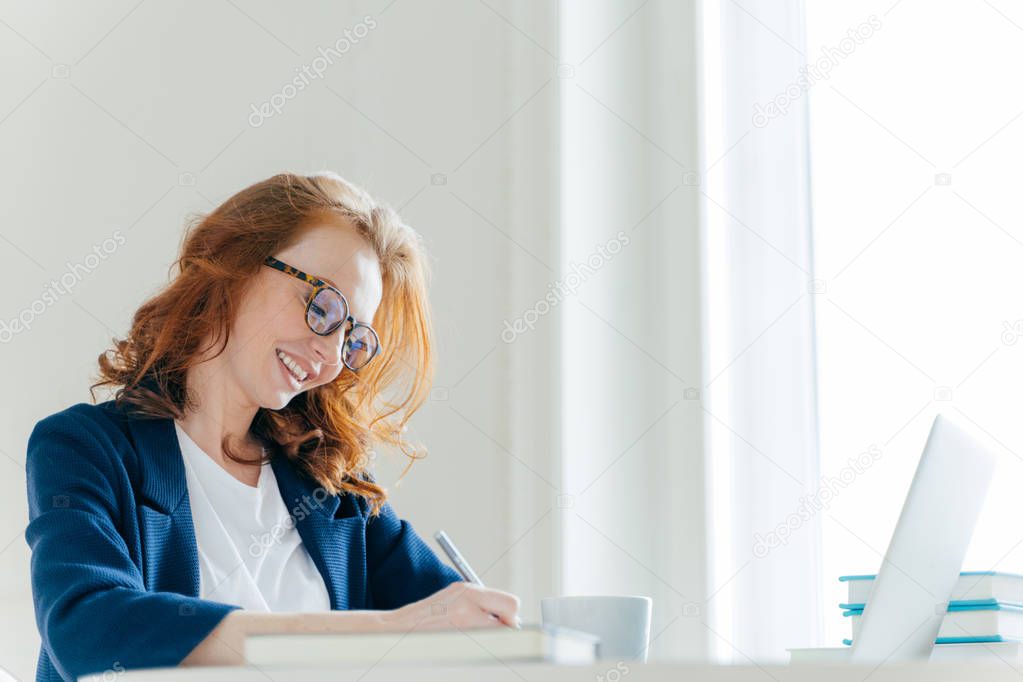 Indoor shot of pleased cheerful famale with ginger hair, signs contract, sits at cabinet in front of laptop computer. Smiling director notes information for financial report, works on laptop computer