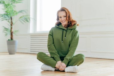 Mutlu zencefil kadın spor eğitmeni yere bacaklar çapraz oturur, yeşil kapüşonlu ve pantolon giyer, beyaz eğitmenler, memnuniyetle gülümsüyor, spor için gitmek için boş odada katta pozlar. Sağlıklı yaşam tarzı