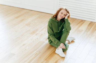 Memnun dinlendirici kadın nazik bir gülümseme, makyaj, spor giyer, beyaz spor ayakkabı, laminat zemin üzerinde pozlar, yoga veya egzersiz egzersizleri uygulamaları, mutluluk ifade eder. İnsanlar ve yaşam tarzı kavramı