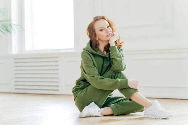 Staying in good shape. Pleasant looking red haired female gymnast with wavy hair, sits on floor in fitness hall, has flexible body, rests after doing yoga exercises, looks confidently at camera