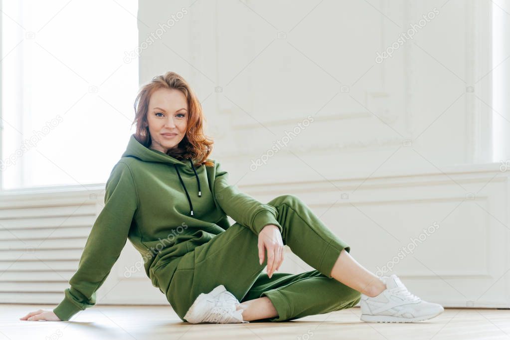 Sideways shot of satisfied redhead woman has makeup, wears tracksuit, white spotshoes, rests on floor, does stretching exercises, is fond of sport. People, flexibility and motivation concept