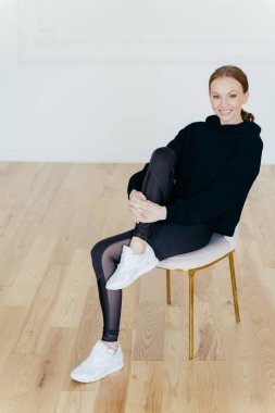 Zencefil penye saçları ile Memnun kadın, hoş bir gülümseme vardır, bacak yükseltir ve diz üzerinde el tutar, stüdyoda sandalyede pozlar, aerobik sonra dinlenme vardır, siyah spor kostüm ve spor ayakkabı giyer. Motivasyon