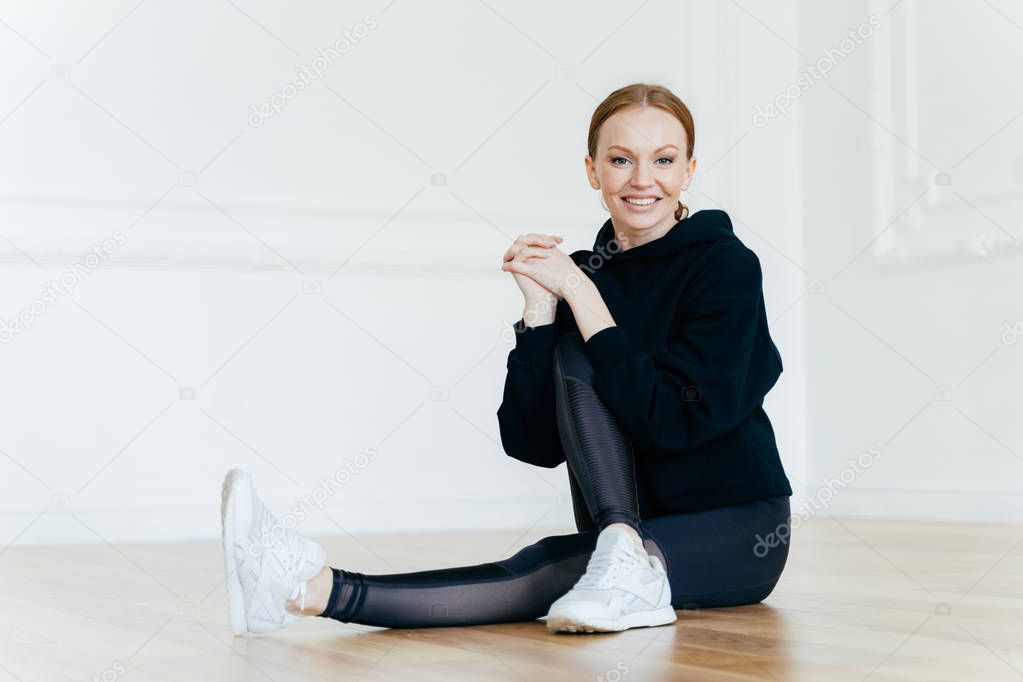 People, sport, recreation concept. Delighted red haired female has positive smile, sits on floor in empty room, leads healthy lifestyle, does fitness exercises, wears hoody, leggings and sneakers