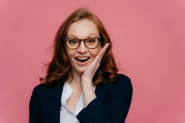 Mutlu kadın entreprenuer yanak hafifçe dokunur, geniş gülümsüyor, şeffaf gözlük giyer, zencefil saçlı, resmi giyim giymiş, başarı elde etmek için memnun, pembe arka plan üzerinde izole
