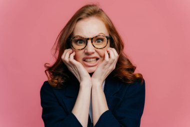 Olumsuz duygu kavramı. Memnun redhead müreffeh iş kadını tahriş dişleri sıkıyor, çene altında ellerini tutar, şeffaf gözlük ve siyah resmi kıyafet giyer, pembe duvarda izole