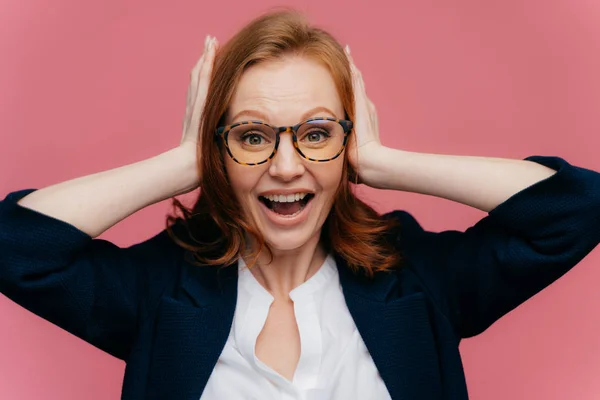 Photo of positive redhead female office worker keeps palms on ears, looks happily at camera, wears formal outfit, isolated over pink background. People, positive emotions and facial expressions