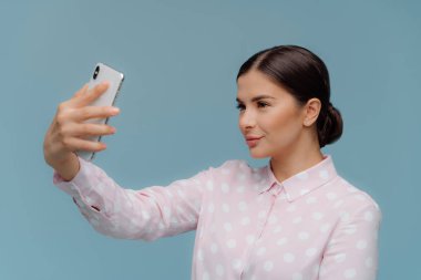 Koyu saçlı çekici zarif kadın öğretmen, modern hücresel el uzanır, selfie portre yapar, şık polka nokta gömlek giyer, mavi arka plan karşı duruyor. İnsanlar, teknoloji kavramı