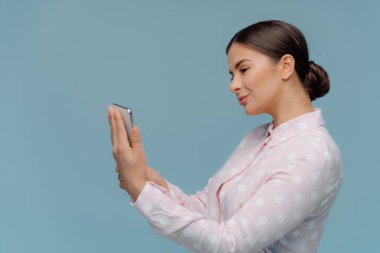 Güzel genç kadın yan yana duruyor, koyu saçlı topuz taranmış, modern akıllı telefon tutar, online sohbetler, sosyal ağlarda paylaşmak için güzel bir resim seçer, mavi arka plan üzerinde izole.