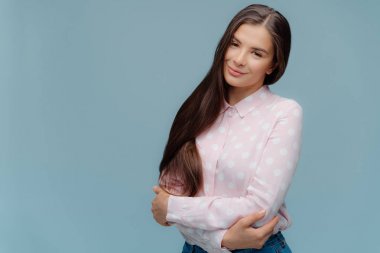 Uzun koyu lüks saçlı iyi görünümlü kadın Portresi elleri çapraz tutar, kamera mutlu görünüyor, utangaç hissediyor, polka nokta gömlek giyer, sloganı için kopya alanı ile mavi arka plan üzerinde modeller