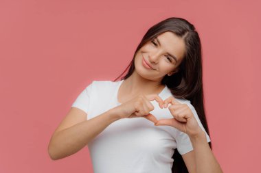 Gülümseyen memnun esmer kadın model şekiller kalp işareti, erkek arkadaşı aşk ifade eder, rahat beyaz t shirt giymiş, pembe arka plan üzerinde izole. Sen benim sevgilimsin! Vücut dili kavramı