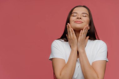 Hoş duygular. Mutlu esmer kadın spa prosedürleri sonra cilt yumuşaklık sahiptir, çene dokunur, gözleri kapalı tutar, baş kaldırır, gözleri kapatır, zevk hissediyor, beyaz t shirt giyer, izole