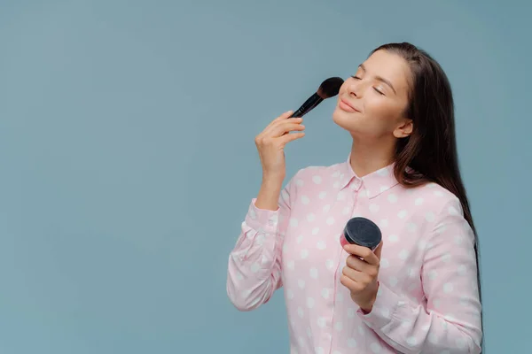 Satisfied lady applyes foundation with cosmetic brush, does professional makeup, wears elegant stylish shirt, models over blue background with copy space for your promotion. Beauty and cosmetology