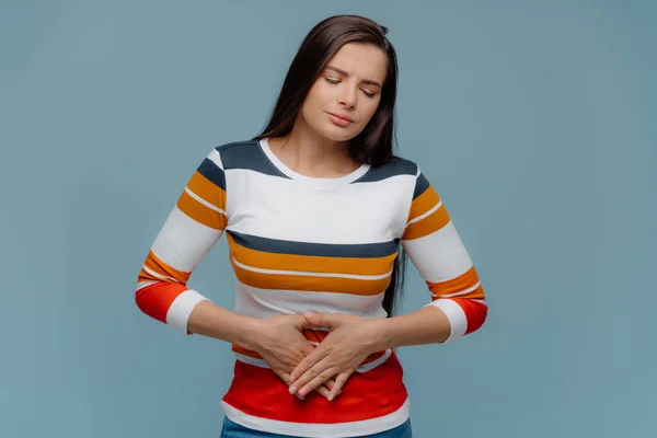 Upset brunette lady feels unwell, suffer from stomachache, feels discomfort in belly, has dissatisfied facial expression, dressed in white jumper with colored stripes, isolated on blue background