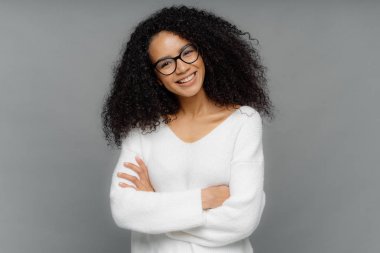 Dost görünümlü Afro-Amerikan kadın yarım uzunlukta çekim baş eğir, koyu gevrek gür saçları vardır, göğüs üzerinde katlanmış kollarını tutar, rahat beyaz kazak giyer, stüdyoda photoshoot sahiptir. Duyguları