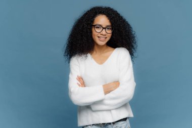 Mutlu görünümlü kıvırcık kadın Portresi göğüs üzerinde katlanmış kollarını tutar, kendinden emin yüz ifadesi vardır, yüzünde charmin gülümseme, beyaz kazak ve kot giyer, mavi arka plan üzerinde duruyor.