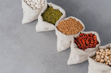 Gri arka plan üzerinde keten bez torbalarda çeşitli glutensiz tahıllar. Çuvallar böbrek fasulyesi, mercimek, karabuğday, goji ve garbanzo ile dolu. Sağlıklı beslenme için tahıllar