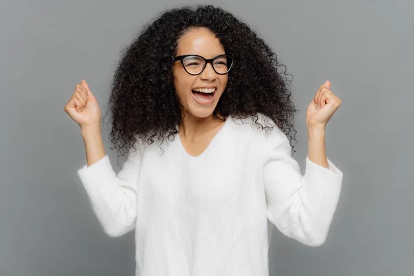 Photo en studio d'une femme à la peau foncée triomphante serrant les poings, souriant joyeusement, célébrant le succès, atteignant l'objectif, souriant largement, vêtue de façon désinvolte, isolée sur fond gris. Concept de victoire — Photo