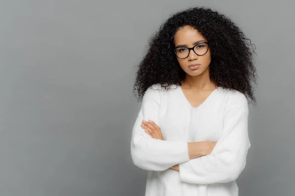 Self confident dark skinned ethnic female keeps hands crossed over chest, looks seriously at camera, wears optical spectacles and white sweater, stands against grey background with empty space — Stock Photo, Image