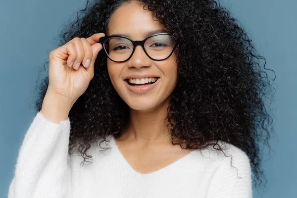 Обрізаний знімок красивої жінки носить оптичні окуляри, посміхається широко, показує білі ідеальні зуби, здорову темну шкіру, одягнену в повсякденний джемпер, ізольовані на синьому фоні. концепція жіночності — стокове фото