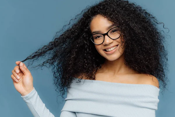 Plan horizontal d'une belle femme afro-américaine souriante tient les pointes de ses cheveux bouclés, porte un pull bleu clair à la mode, des lunettes transparentes, des modèles sur fond bleu. Regarde mes boucles. — Photo