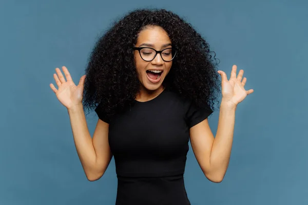 Slim femme à la peau foncée surémotive lève les mains, ouvre la bouche, gestes activement des émotions positives, concentré vers le bas, vêtu d'un t-shirt noir décontracté, isolé sur fond bleu. Concept d'ethnicité — Photo