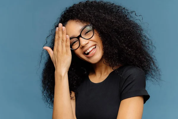 Happy Afro American woman touches forehead, tilts head, smiles happily at camera, has fun indoor, wears optical spectacles and casual black t shirt, isolated over blue background. Emotions concept