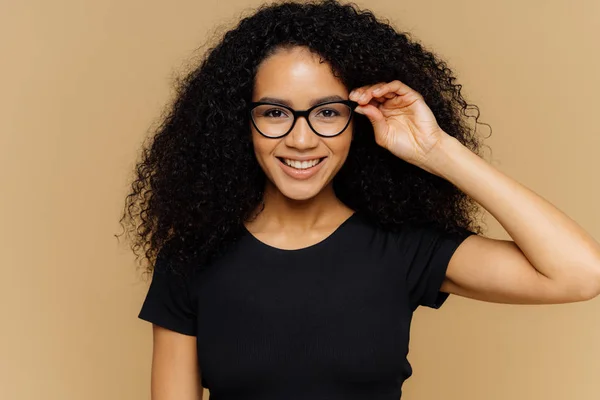 Härlig ung afrikansk amerikansk kvinna flinar vid kameran, håller handen på kanten av glasögon, glad att höra goda nyheter, klädd i casual svart t-shirt, isolerad över brun bakgrund. Happiness Concept — Stockfoto