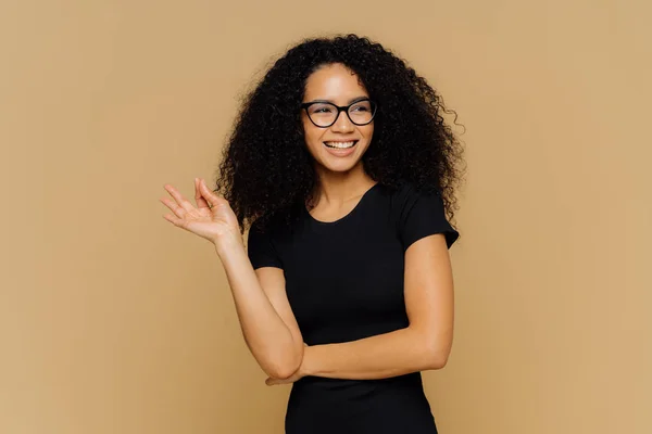 Усміхнена жінка з афро стрижка, робить добре жест, виглядає щасливо в сторону, погоджується з мишей пропозиції, носить оптичні окуляри і випадкові чорної футболки, має гарну фігуру, ізольовані на коричневому фоні — стокове фото