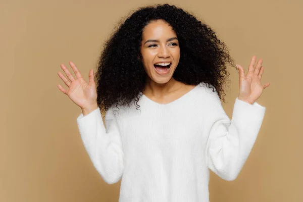 Heureuse femme surémotive surprise par une pertinence agréable, lève les mains et montre les paumes, se concentre de côté, porte un pull blanc décontracté, des modèles sur fond brun. Émotions et sentiments humains positifs — Photo