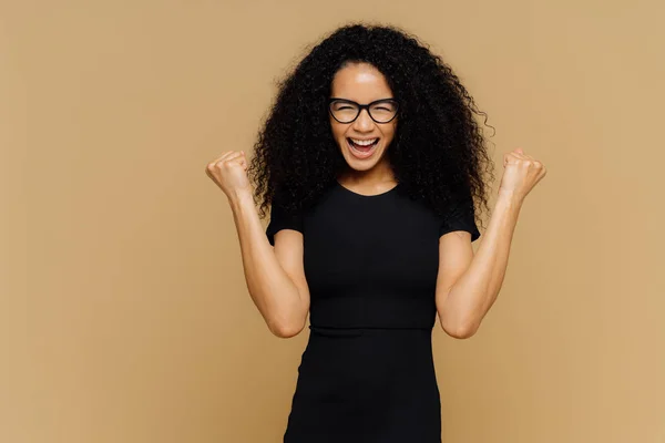 Studio shot of overjoyed cheering woman with curly hair, lève les poings serrés, célèbre le succès et le triomphe, vêtu d'un t-shirt noir décontracté, lunettes, se dresse contre un mur brun avec un espace vide — Photo