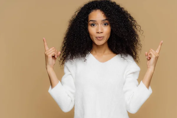 Photo of confident woman has bushy Afro hairstyle, points both index fingers above, dressed in white jumper, stands against brown background, advertises some item, shows direction. Come upstairs — Stock Photo, Image