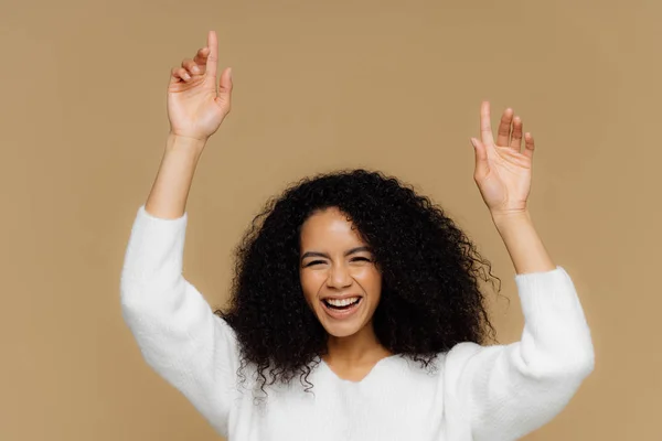 Горизонтальний знімок прекрасної темношкірої жіночої моделі має зубну посмішку, піднімає руки, вказує вгору, відчуває себе задоволеним, має кучеряве волосся, носить білий светр, ізольований на коричневому тлі. Жінка розважається — стокове фото