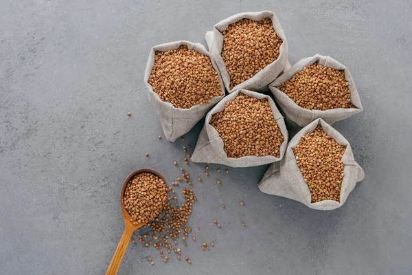 Top view of brown buckwheat in sacks and wooden spoon over grey background. Healthy diet concept. Buckwheat groats. Biologically grown organic plant — Stock Photo, Image