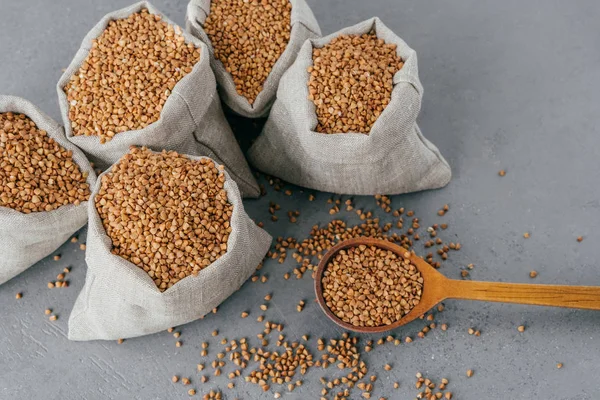 Vista superior de cinco sacos pequeños llenos de trigo sarraceno crudo, cuchara de madera cerca con cereales sin cocer, fondo oscuro. Concepto de comida saludable — Foto de Stock