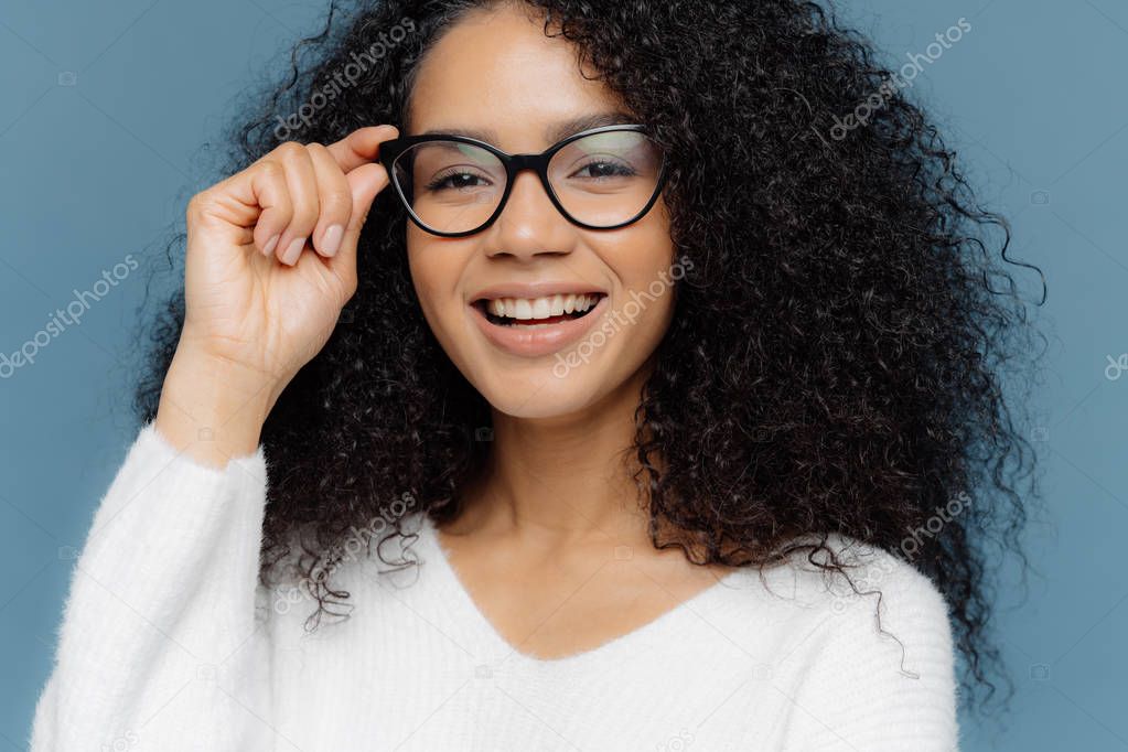 Cropped shot of good looking woman wears optical glasses, smiles broadly, shows white perfect teeth, healthy dark skin, dressed in casual jumper, isolated over blue background. Feminity concept
