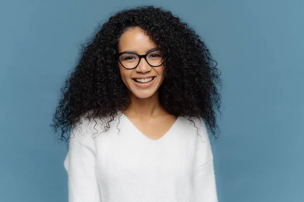 Headshot av härlig lockig kvinna är på gott humör, ler brett, uttrycker fina känslor, bär genomskinliga glasögon och vita Jumper, isolerade över Blå bakgrund. Afro hona poserar inomhus — Stockfoto