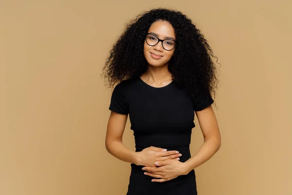Glad afro amerikansk kvinna med lockigt hår, håller händerna på magen, känns bra efter att ha ätit näringsrik läcker middag, ser glatt på kameran. Unga glada kvinnliga upptäcker om graviditet. — Stockfoto