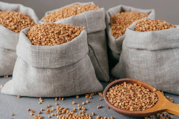 Primer plano de saco hecho de tela de lino lleno de trigo sarraceno marrón seco, cuchara de madera con cereal crudo. Enfoque selectivo — Foto de Stock