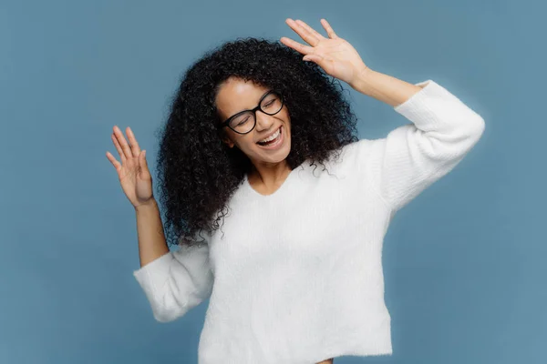 Les gens et le concept de joie. Optimiste ravie Afro American femme lève la main, danse sur la musique forte, être ravi, porte cavalier blanc décontracté, isolé sur fond bleu, a bonne humeur — Photo